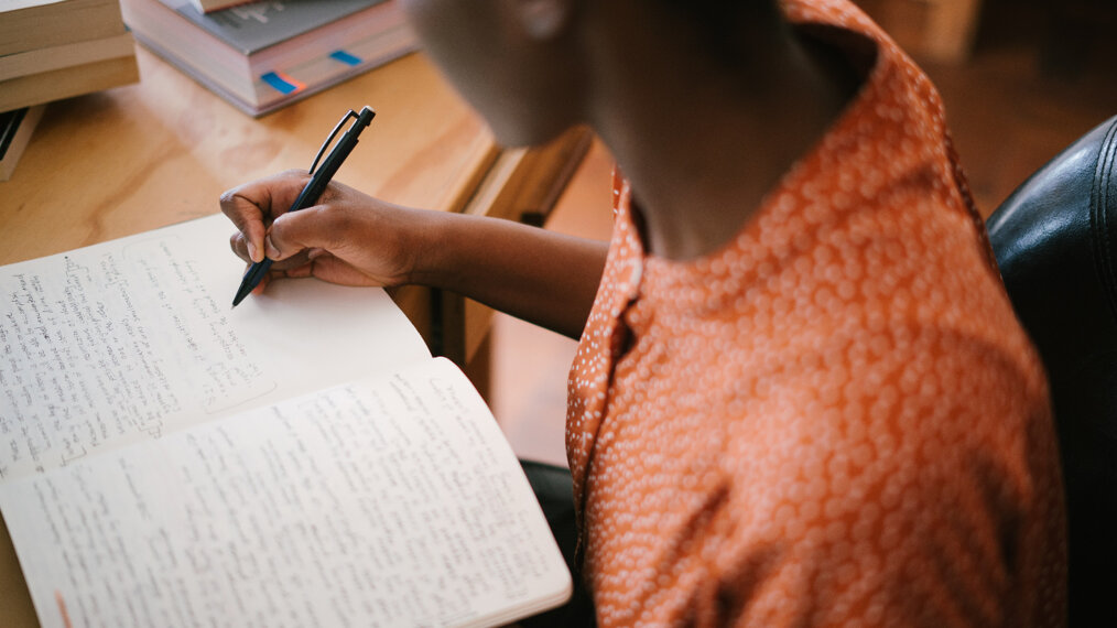 Une personne est assise à un bureau. Elle est face à un cahier et semble écrire dedans.