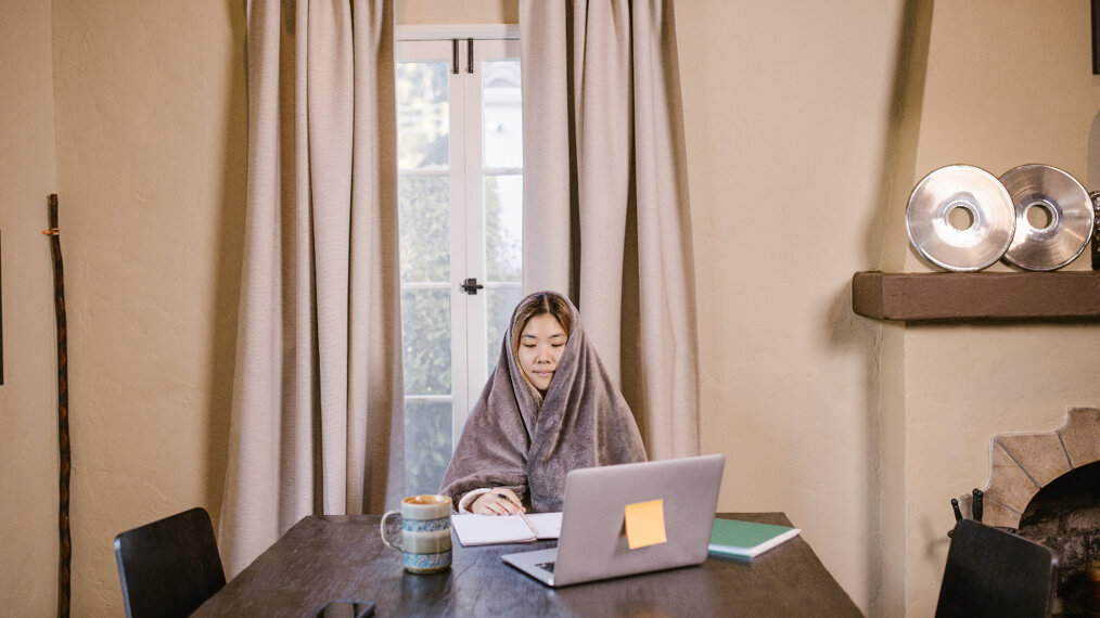 Une jeune fille révise sur un bureau, enroulée dans un plaid et avec une tasse à côté.