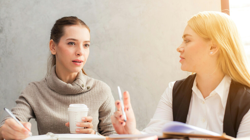 Une jeune fille et une femme discutent. Elles sont assises autout d'une table pleine de cahiers ouverts.